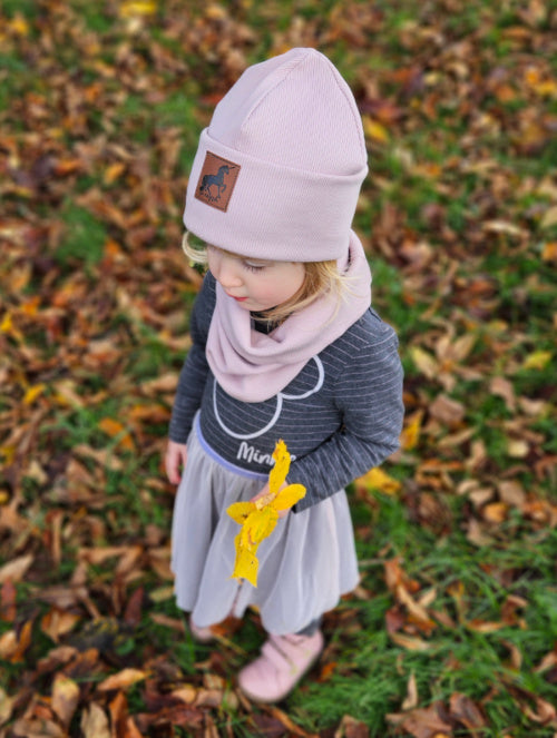 Ein kleines Mädchen trägt ein Set aus Hipster-Beanie und Loop aus Rib-Jersey in der Farbe altrosa. Auf der Mütze befindet sich ein brauner Kunstlederpatch mit einem schwarzen Einhorn.